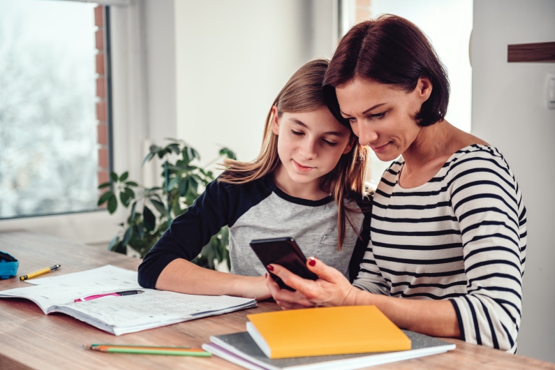 A mother and daughter sit at a table and review a calculator while discussing tips for teaching kids about money.