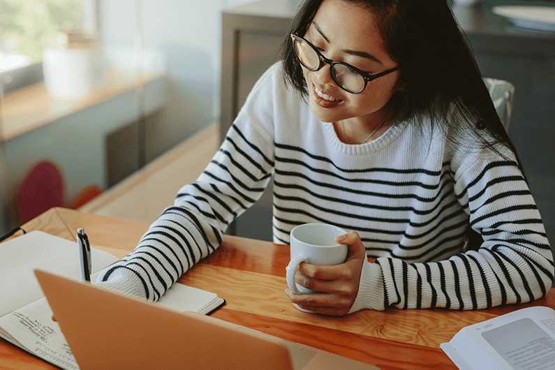 Woman wearing a black and white striped sweater sitting at a desk taking notes while holding a coffee cup.