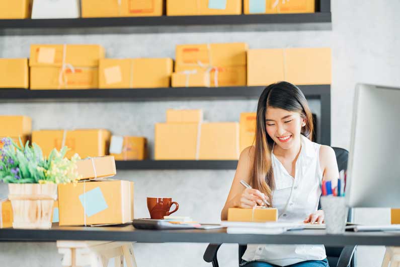 A woman in her mid-20s smiles as she sits at a black table and writes on paper among stacks of yellow shipping boxes.