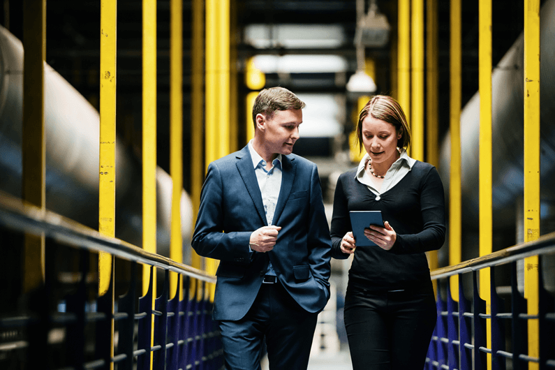 Man and woman walk down a row of IT machinery while looking at a tablet.