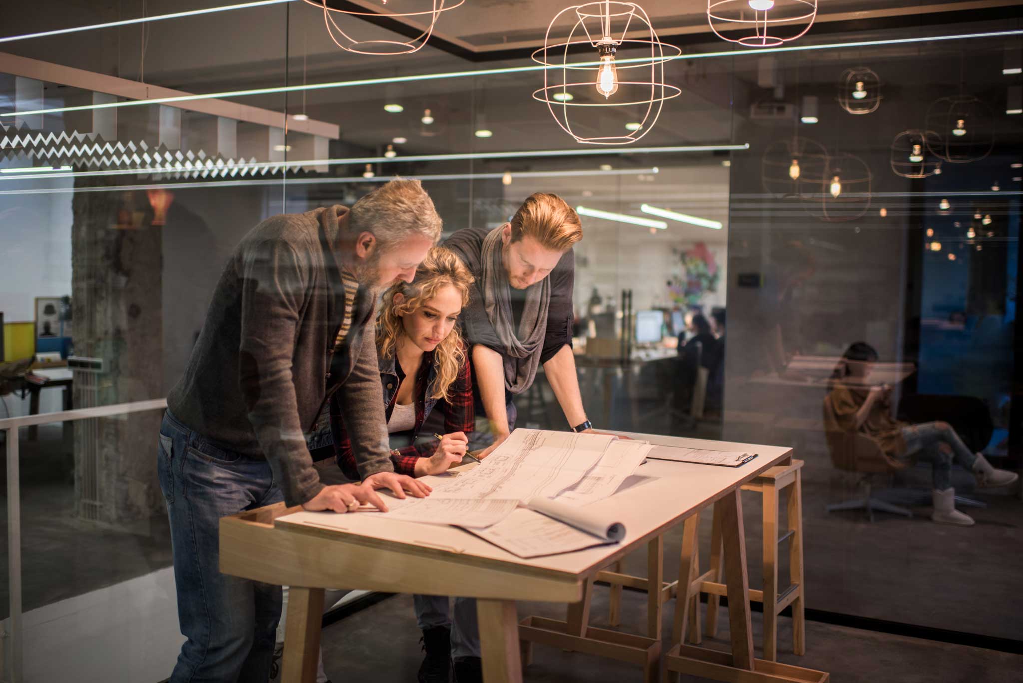 A group of three project managers overlooking printed construction plans in a brightly lit office space.