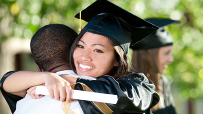 A young woman smiles and hugs her father after receiving a college diploma.