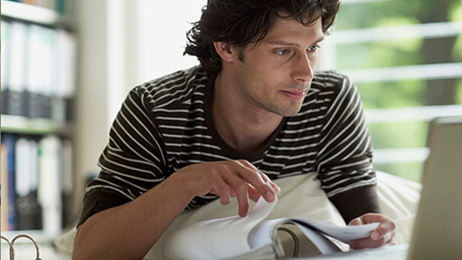 A male looking at the computer with a binder of papers in his hand