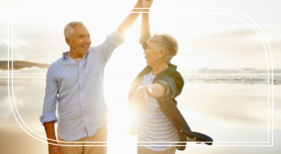 A retired couple dance on the beach during a sunset and smile after enjoying their retirement advisory services.